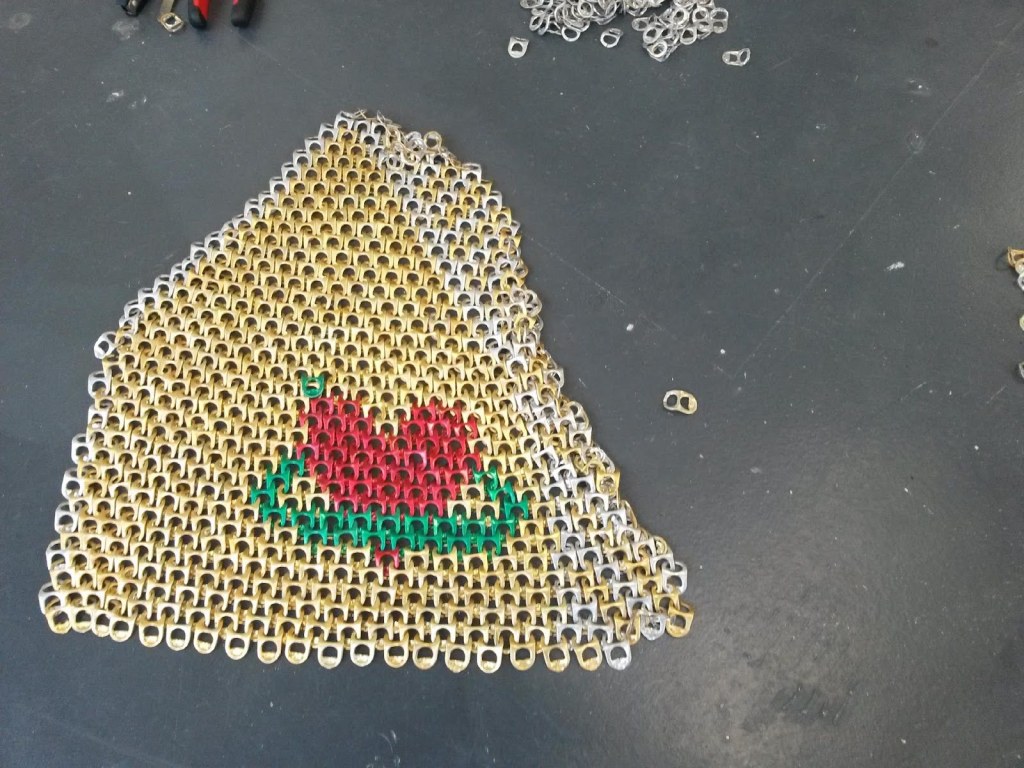 A photograph of a flat object lying on a grey floor. The object is an irregular shaped piece of chainmail like work that is mage from connected gold and silver ring pulls. The chainmail has a motif of a red heart surrounded by a green ring made from the appropriately coloured ring pulls. 